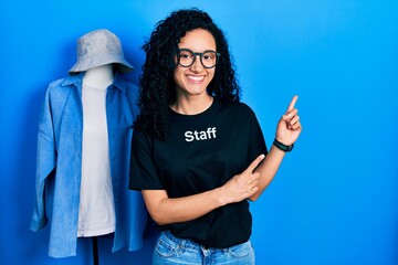 Young hispanic woman with curly hair wearing staff t shirt covering mouth with hand, shocked and...