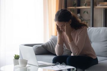 Young woman sit on sofa near laptop takes off glasses touch temples suffers from migraine headache...