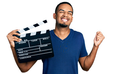 Young african american man holding video film clapboard screaming proud, celebrating victory and...