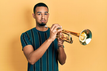 Young african american man playing trumpet puffing cheeks with funny face. mouth inflated with air,...