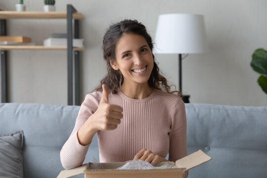 Latin woman sit on sofa at home with opened parcel box feels satisfied by delivered goods show thumbs up gives positive feedback look at camera, trusted transporting company, express delivery concept