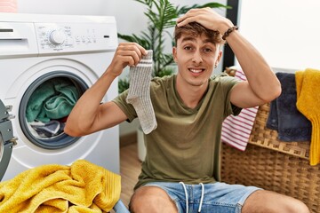 Young caucasian man doing laundry holding sock stressed and frustrated with hand on head, surprised...