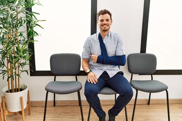 Handsome young man sitting at doctor waiting room with arm injury with a happy and cool smile on face. lucky person.