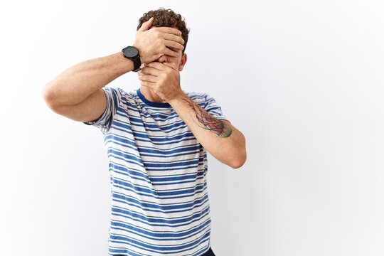 Handsome young man standing over isolated background covering eyes and mouth with hands, surprised and shocked. hiding emotion