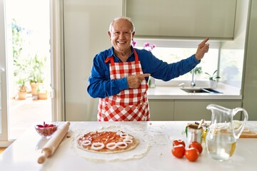 Senior man with grey hair cooking pizza at home kitchen smiling and looking at the camera pointing with two hands and fingers to the side.