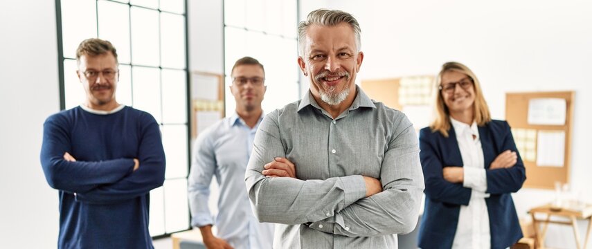 Middle age businessman smiling with arms crossed gesture standing with work partners at the office.