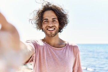 Young hispanic man smiling happy make selfie by the camera standing at the beach.