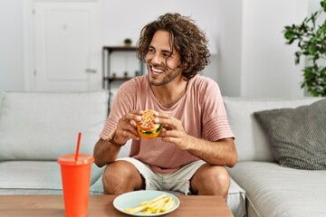 Young hispanic man smiling happy eating classic burger and drinking soda at home.