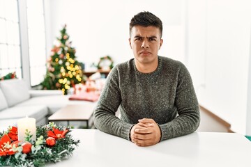 Young hispanic man sitting on the table by christmas tree skeptic and nervous, frowning upset...