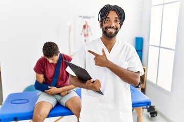 Young hispanic man working at pain recovery clinic with a man with broken arm cheerful with a smile on face pointing with hand and finger up to the side with happy and natural expression