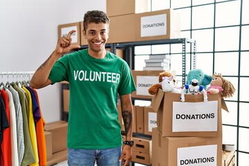 Young handsome hispanic man wearing volunteer t shirt at donations stand smiling and confident gesturing with hand doing small size sign with fingers looking and the camera. measure concept.