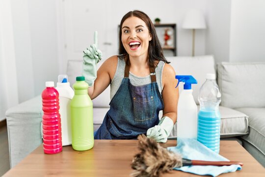 Young brunette woman wearing cleaner apron and gloves cleaning at home pointing finger up with successful idea. exited and happy. number one.