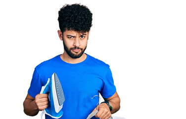 Young arab man with beard holding electric steam iron and white t shirt skeptic and nervous,...