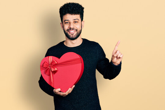 Young arab man with beard holding valentine gift smiling happy pointing with hand and finger to the side