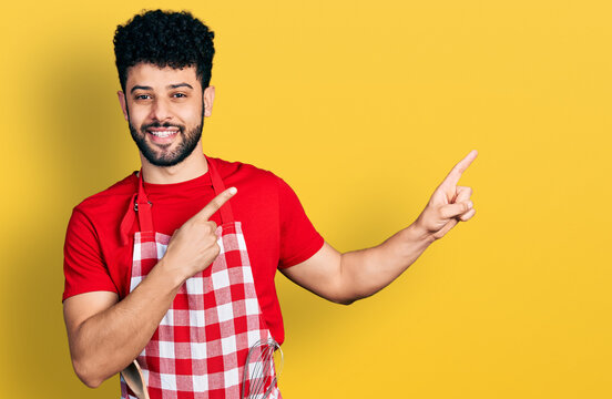 Young arab man with beard wearing cook apron smiling and looking at the camera pointing with two hands and fingers to the side.
