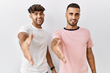 Young gay couple standing over isolated background smiling friendly offering handshake as greeting and welcoming. successful business.