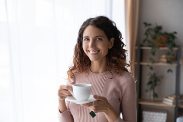 Happy young Latina woman standing in cozy room smile looks at camera holds cup of fresh brewed tea, enjoy no stress calm positive pastime, new day drink favourite beverage. Leisure, lifestyle concept