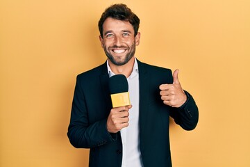 Handsome man with beard holding reporter microphone smiling happy and positive, thumb up doing...