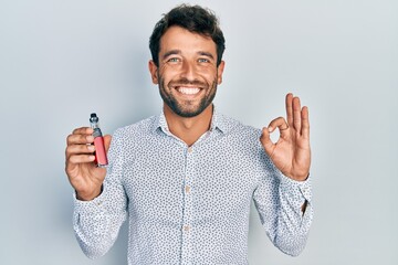 Handsome man with beard football reporter holding electronic cigarette doing ok sign with fingers,...