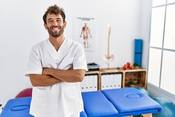 Young handsome physiotherapist man working at pain recovery clinic happy face smiling with crossed...
