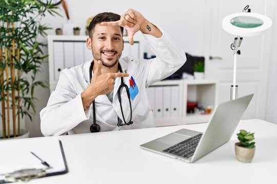 Young doctor working at the clinic using computer laptop smiling making frame with hands and fingers with happy face. creativity and photography concept.