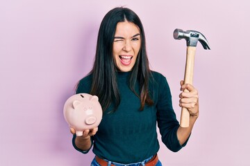 Young brunette woman holding piggy bank and hammer winking looking at the camera with sexy expression, cheerful and happy face.