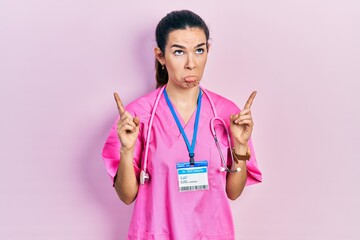 Young brunette woman wearing doctor uniform and stethoscope pointing up looking sad and upset,...