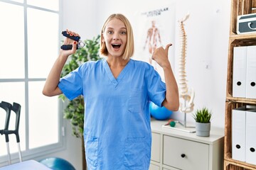 Young caucasian physiotherapist woman holding hand grip to train muscle pointing thumb up to the side smiling happy with open mouth