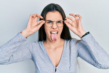 Young brunette teenager wearing glasses sticking tongue out happy with funny expression.