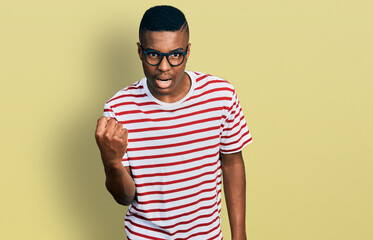 Young african american man wearing casual t shirt and glasses angry and mad raising fist frustrated and furious while shouting with anger. rage and aggressive concept.