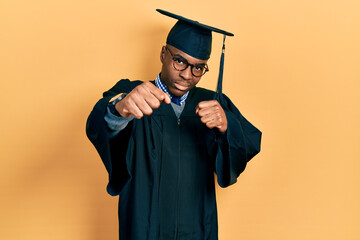 Young african american man wearing graduation cap and ceremony robe punching fist to fight,...