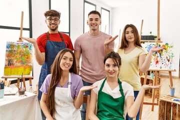 Group of five hispanic artists at art studio smiling cheerful presenting and pointing with palm of hand looking at the camera.