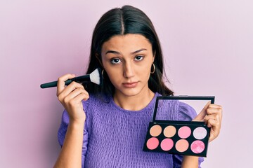 Young hispanic woman holding makeup brush and blush skeptic and nervous, frowning upset because of...