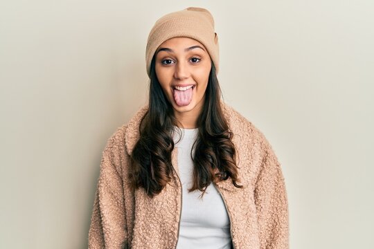 Young hispanic woman wearing wool sweater and winter hat sticking tongue out happy with funny expression. emotion concept.