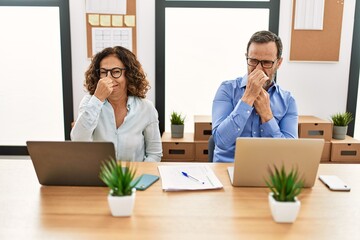 Middle age hispanic woman and man sitting with laptop at the office smelling something stinky and disgusting, intolerable smell, holding breath with fingers on nose. bad smell - obrazy, fototapety, plakaty
