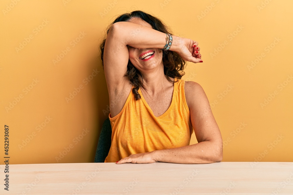 Poster beautiful middle age woman wearing casual clothes sitting on the table covering eyes with arm smilin