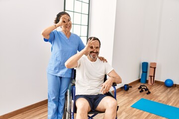 Hispanic middle age man sitting on wheelchair and nurse at rehabilitation clinic smiling happy doing ok sign with hand on eye looking through fingers