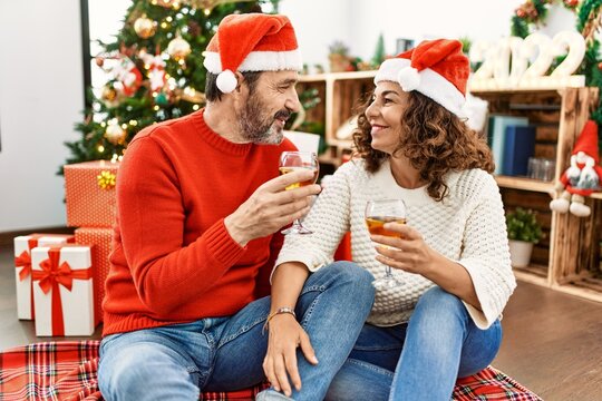 Middle age hispanic couple wearing christmas hat. Sitting on the floor toasting with champagne at home.