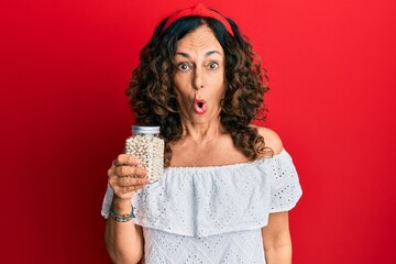 Middle age hispanic woman holding jar of beans scared and amazed with open mouth for surprise, disbelief face