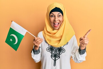 Middle age hispanic woman wearing hijab holding pakistan flag smiling happy pointing with hand and finger to the side - obrazy, fototapety, plakaty