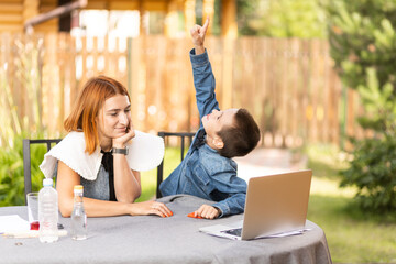 Mom and boy schoolboy are engaged in lessons through a laptop at home in the garden. Online classes for children. Boy shows up to the sky