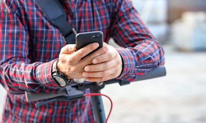 Young man wearing shirt leaning on electric scooter handlebar, holding mobile smartphone in his hands, closeup detail