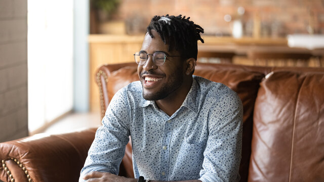 Happy stylish Afro American hipster guy portrait. Young Black man, student, self employed business owner with trendy haircut, glasses, toothy smile sitting on leather sofa at home. Banner, head shot