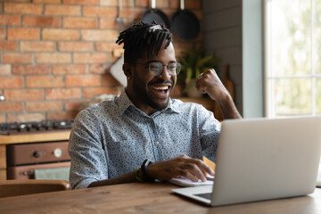 Excited African millennial guy using laptop at home, receiving good news from video talk call, online chat, email, feeling, laughing, shouting. Student celebrating learning success, passed exam