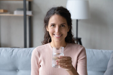 Latin young beautiful woman sit on sofa smile looks at camera holding glass of natural still fresh...
