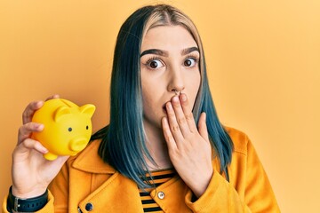 Young modern girl holding yellow piggy bank covering mouth with hand, shocked and afraid for...