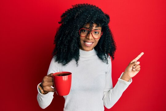 African american woman with afro hair holding coffee smiling happy pointing with hand and finger to the side