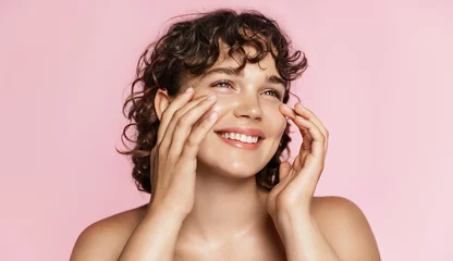 Foto op Canvas Beautiful natural girl smiling, rubbing her face with facial cleanser for glowing healthy skin, looking happy. Curly young woman showering, using skincare hydrating products, pink background © Liubov Levytska