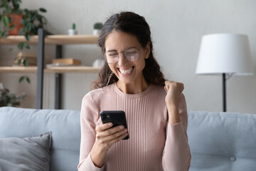 Fototapeta premium Attractive happy Latina woman in glasses sit on sofa at home holding smartphone read sms notification great news looking excited overjoyed cant believe in luck enjoy moment of victory. Success concept