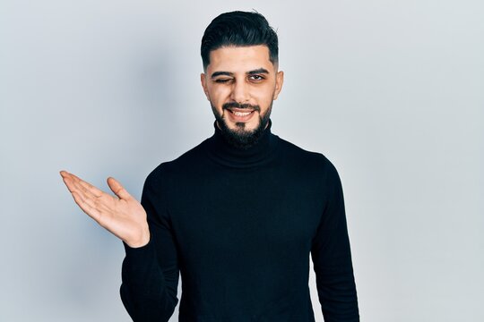 Handsome man with beard presenting with open palm winking looking at the camera with sexy expression, cheerful and happy face.